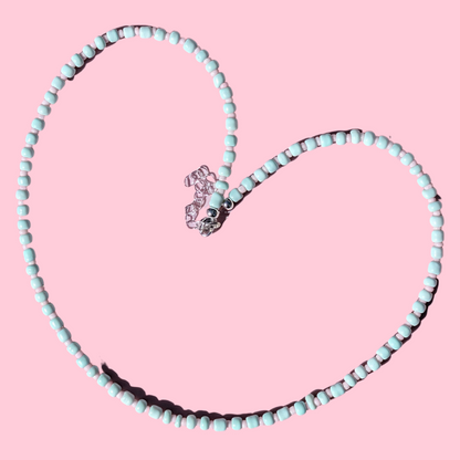 A heart-shaped beaded necklace made of light blue-green beads with a silver-toned lobster clasp, displayed on a solid pink background.