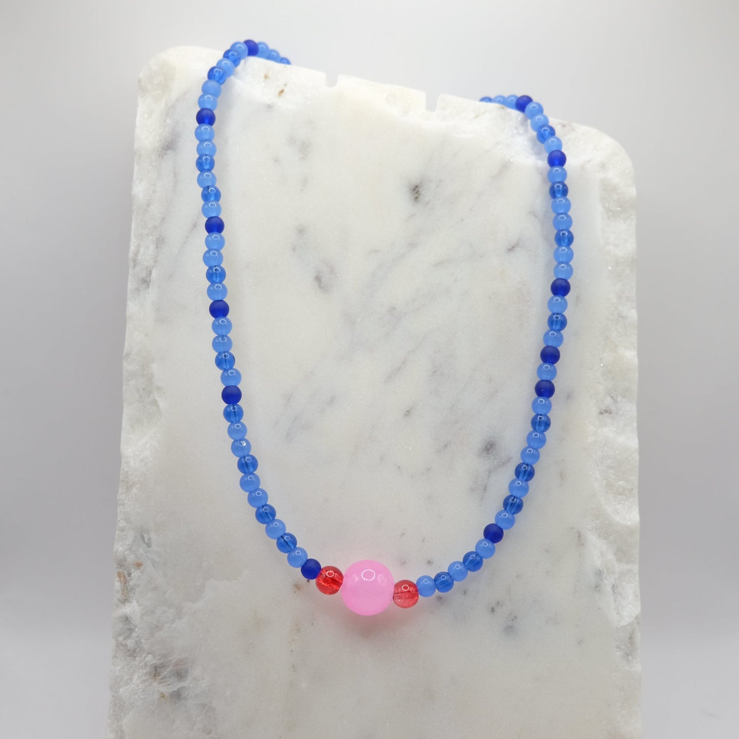 A beaded necklace composed of 4mm blue glass beads - featuring a central pink glass bead with two red glass beads, shown on a white marble background.