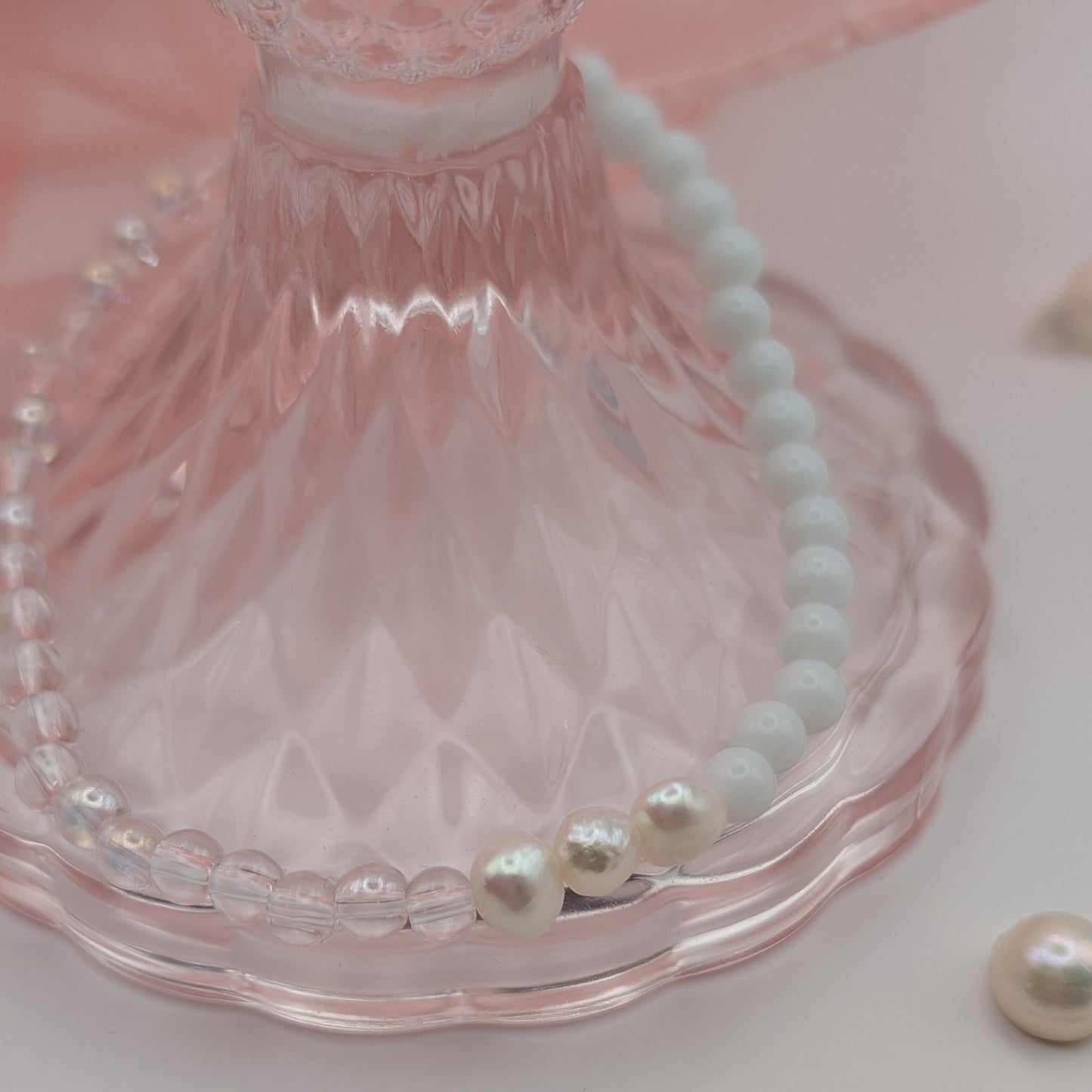 Pearl bracelet on a pink glass stand with scattered pearls on a light pink background