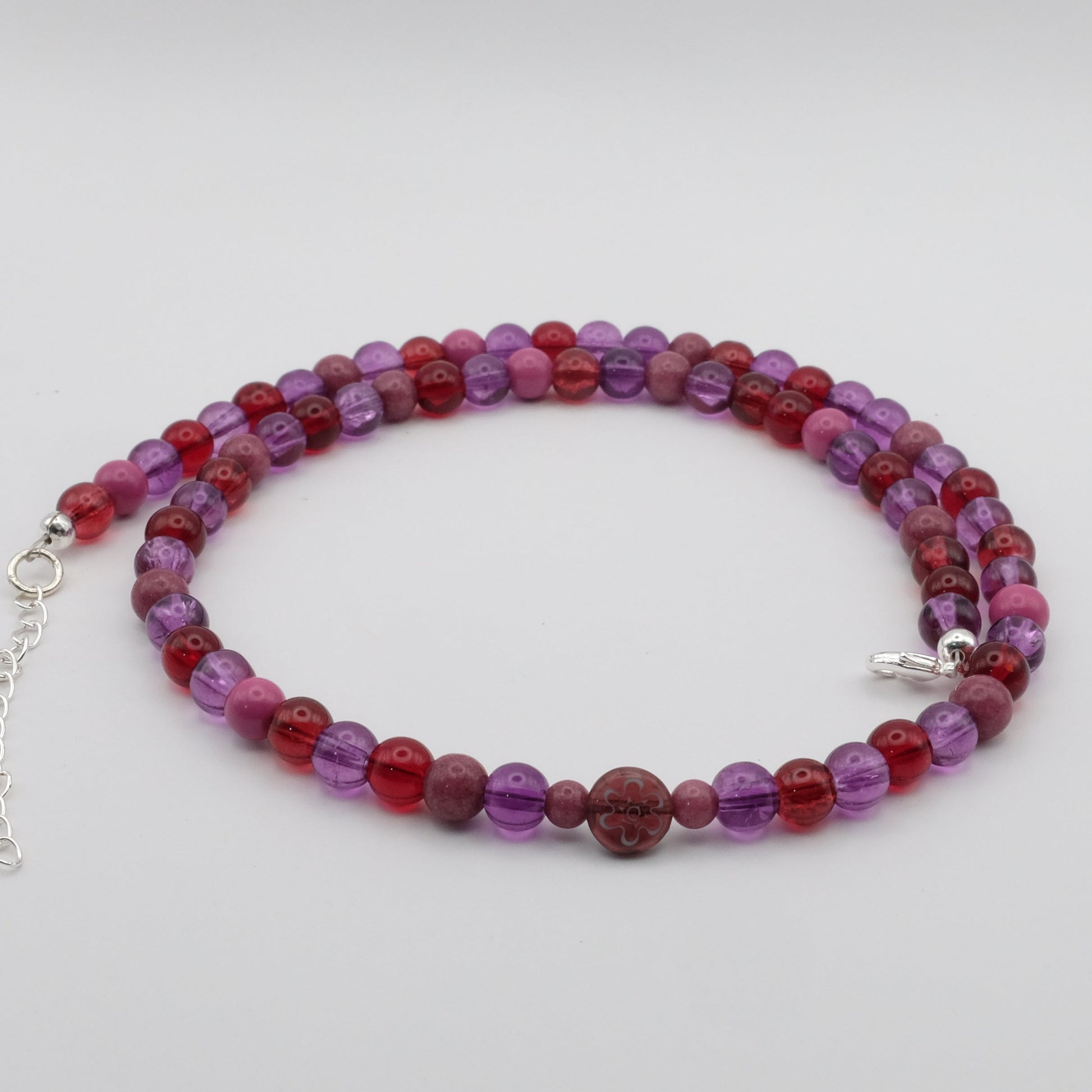 A handmade beaded necklace composed of 6mm glass beads in red, purple and plum tones, featuring a round floral glass bead centrepiece, displayed on a white background.