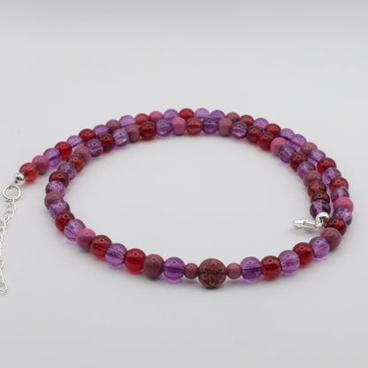 A handmade beaded necklace composed of 6mm glass beads in red, purple and plum tones, featuring a round floral glass bead centrepiece, displayed on a white background.