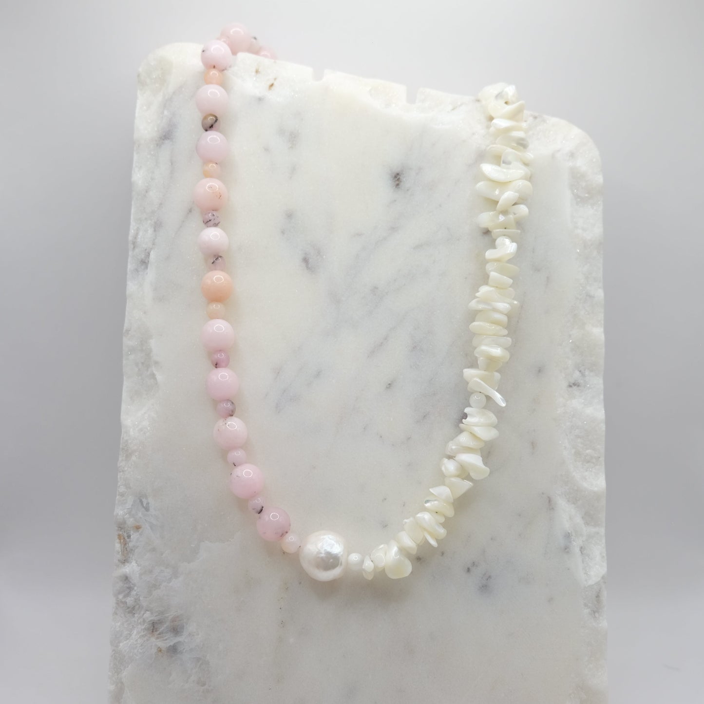 A beaded necklace composed of pink opal beads and mother of pearl chips - featuring a large freshwater pearl nugget at the centre, shown on a white marble background.