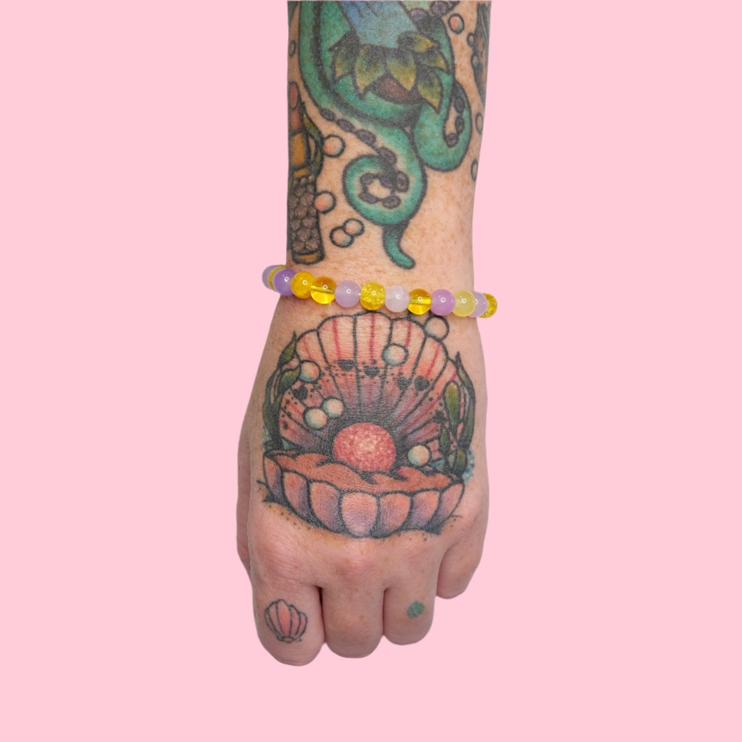 A tattooed arm with octopus and seashell designs wearing an elastic beaded bracelet in purple and yellow hues, set against a solid pink background.