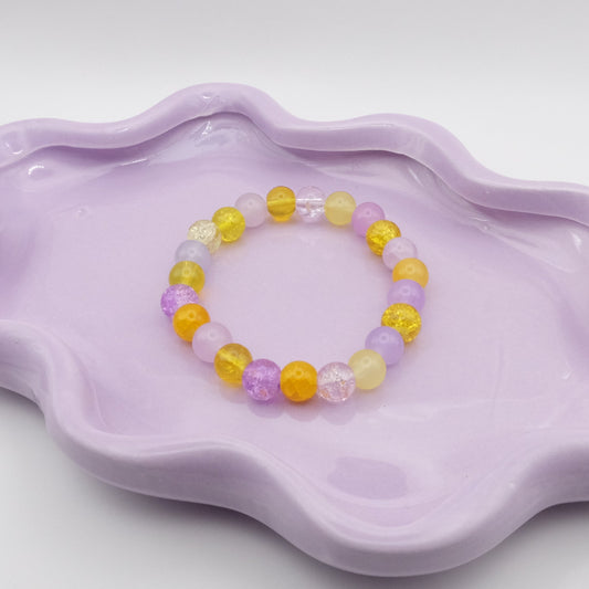 An elastic beaded bracelet composed of 8mm glass beads in soft purple and yellow hues, in varying finishes, displayed on a wavy lavender ceramic dish against a light background.