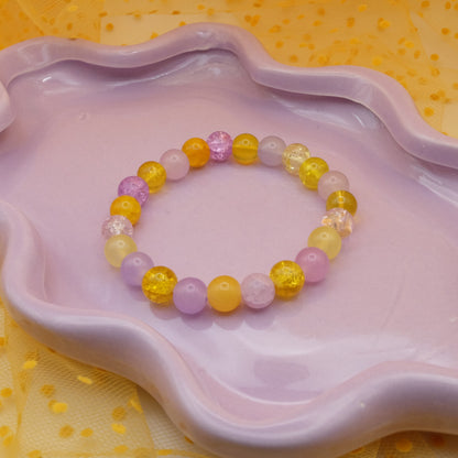 An elastic beaded bracelet from 8mm glass beads in purples and yellows with different finishes, placed on a wavy lavender dish against a yellow polka-dot tulle backdrop.