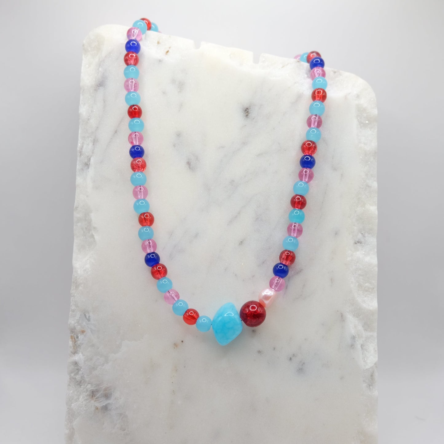 A handmade beaded necklace featuring 6mm glass beads in pink, red and blue, with a dyed blue jade bead, red crackle glass bead and pink freshwater pearl, on a marble background.