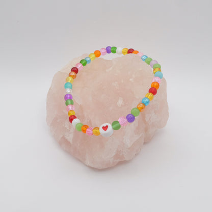 An elastic beaded bracelet composed of colourful 4mm glass beads in red,  orange, yellow, green, aqua, blue,  purple and pink with white and red acrylic love heart bead, displayed on a pale pink ring holder.