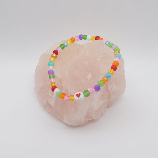 An elastic beaded bracelet composed of colourful 4mm glass beads in red,  orange, yellow, green, aqua, blue,  purple and pink with white and red acrylic love heart bead, displayed on a pale pink ring holder.