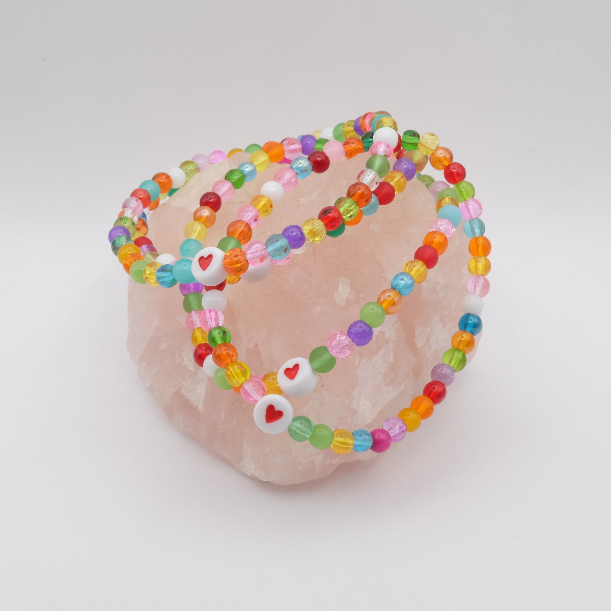 A group of elastic beaded bracelets composed of colourful 4mm glass beads in red, orange, yellow, green, aqua, blue, purple and pink with white and red acrylic love heart bead, displayed on a pale pink ring holder.