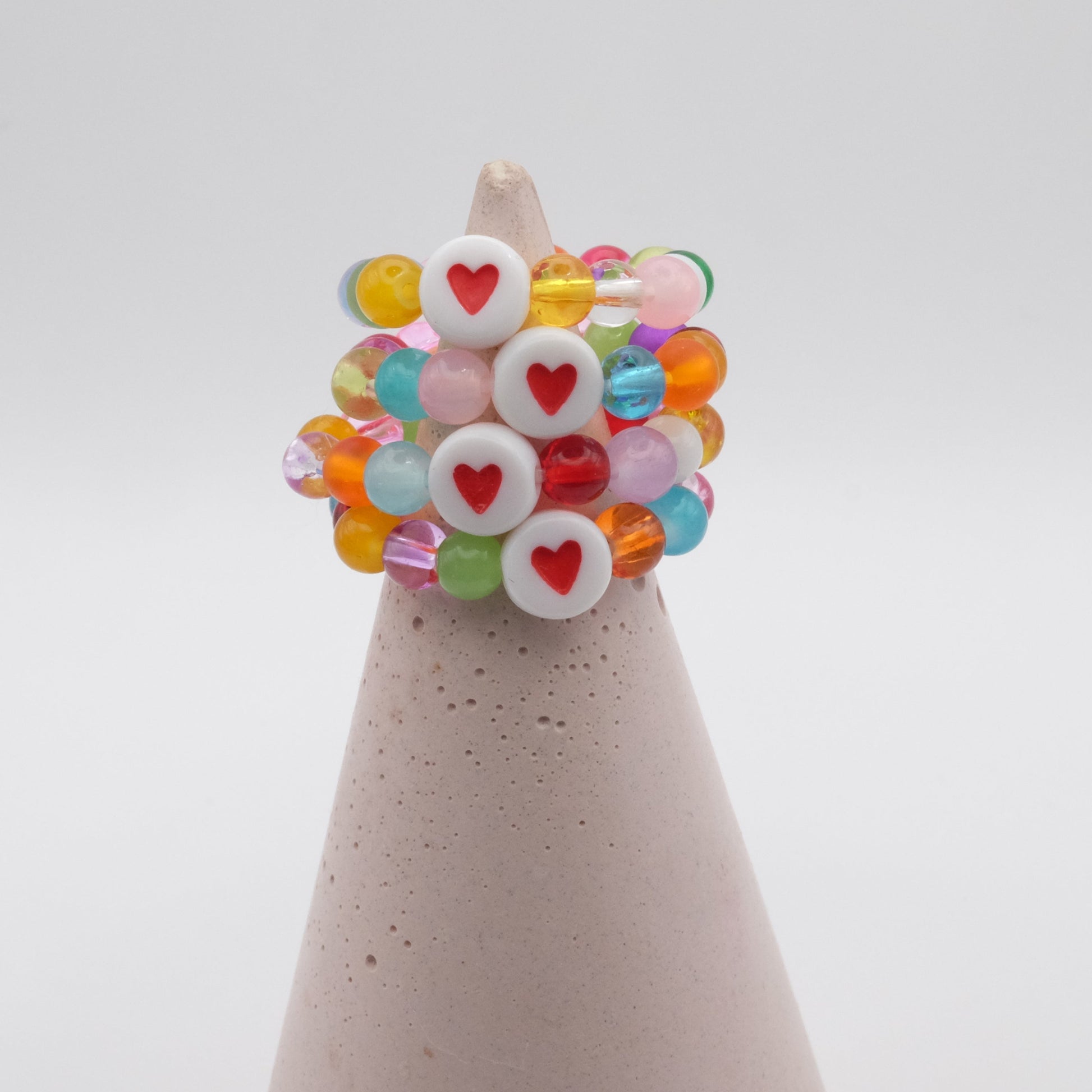 An group of elastic beaded rings composed of colourful 4mm glass beads in red,  orange, yellow, green, aqua, blue,  purple and pink with white and red acrylic love heart bead, displayed on a pale pink ring holder.