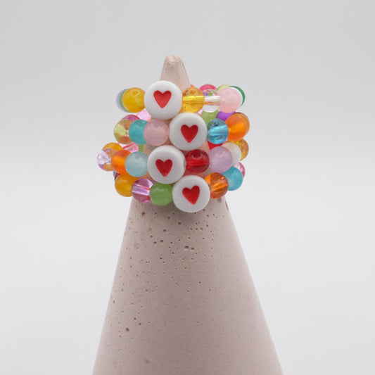 An group of elastic beaded rings composed of colourful 4mm glass beads in red,  orange, yellow, green, aqua, blue,  purple and pink with white and red acrylic love heart bead, displayed on a pale pink ring holder.